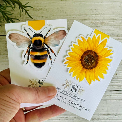 Hand holding a Scattered Seed Co. bookmark with bee and sunflower designs on a wooden surface - The Book Keeper & Co