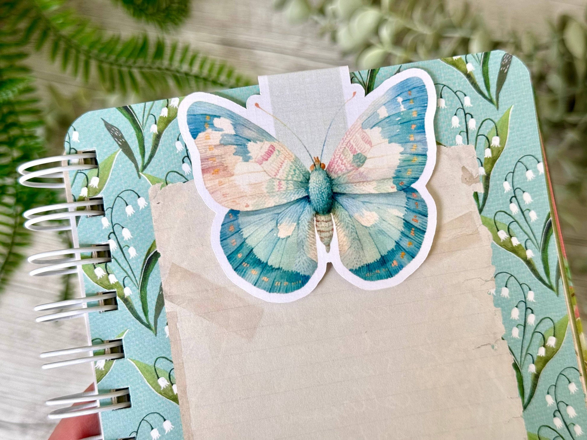 Notebook with a decorative butterfly bookmark  on a green leafy background. - the book keeper and Co