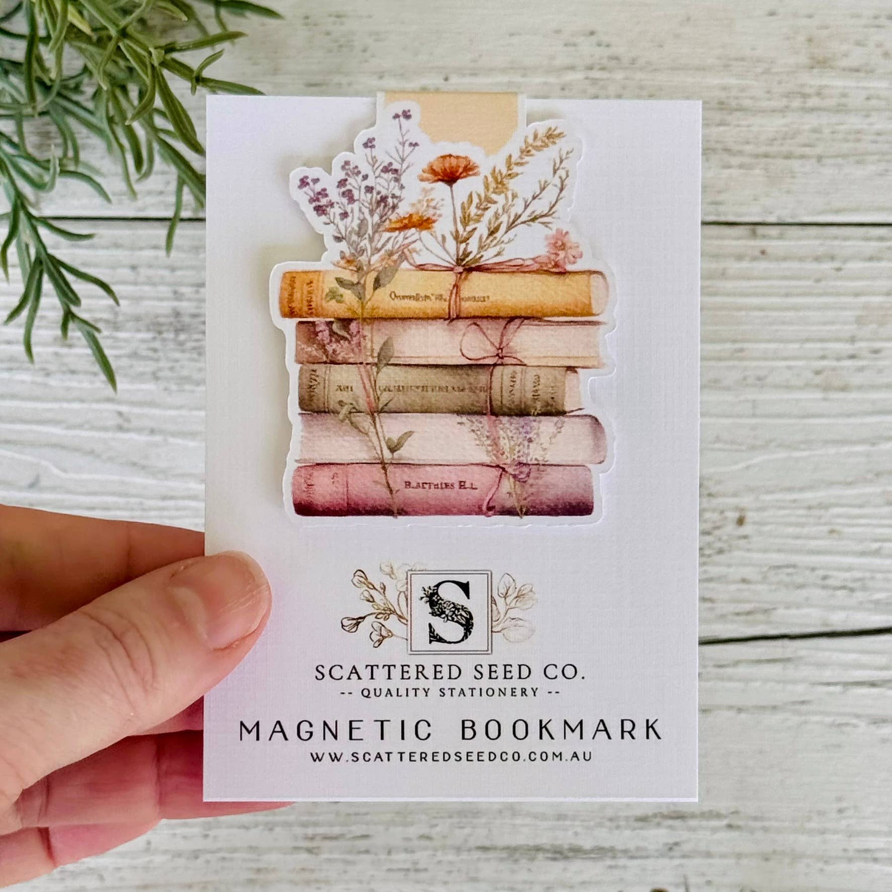 Magnetic bookmark with stack of books and flowers on a card held by a hand, with 'Scattered Seed Co.' branding - The Book Keeper and co