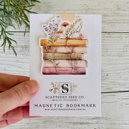 Magnetic bookmark with stack of books and flowers on a card held by a hand, with 'Scattered Seed Co.' branding - The Book Keeper and co